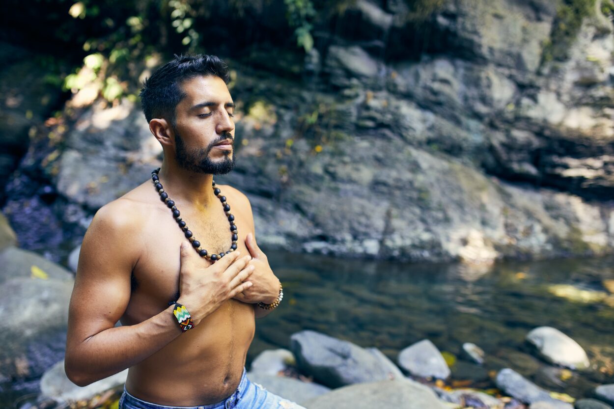Hombre con las tetas al aire meditando cerca del agua.